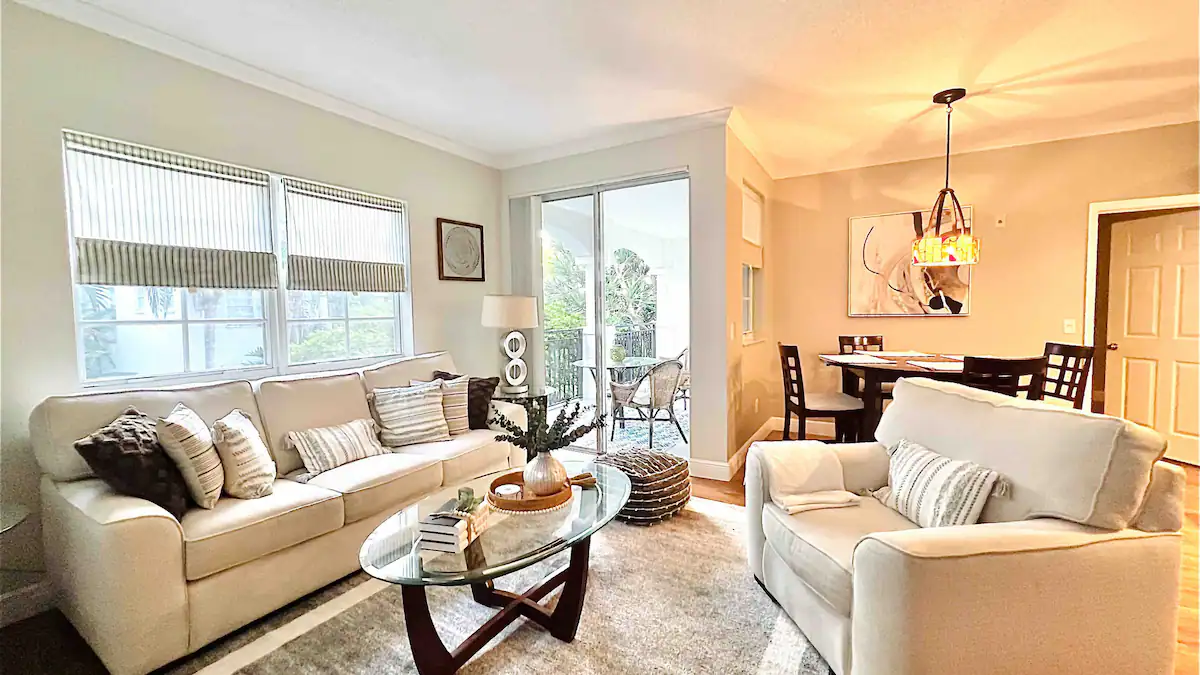 Living room with cream sofas, a glass coffee table, and a chandelier-lit dining area in the Aventura one-bedroom apartment