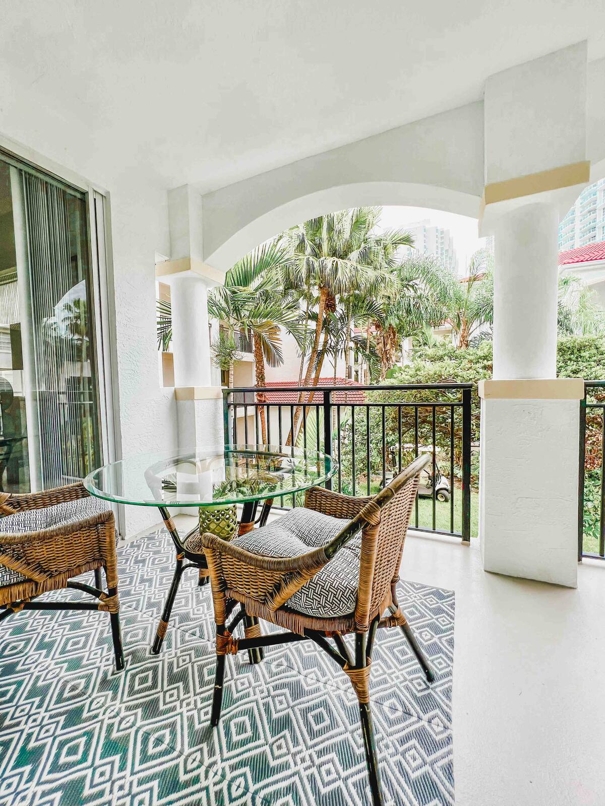 Private balcony with wicker seating and a glass-top table overlooking the tropical courtyard at Village By The Bay